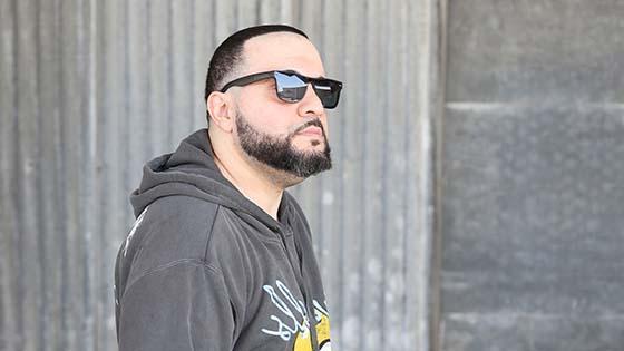 Full Sail grad George Doman wearing black sunglasses and a grey pullover hoodie with a yellow and light blue design, standing in front of a large stone wall while looking off into the distance.